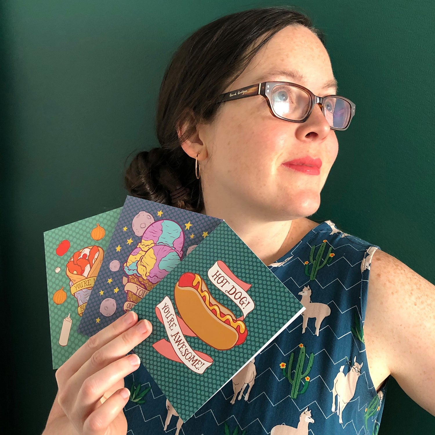 A white woman with dark hair and glasses holds 3 greeting cards. She stands against a dark green wall.