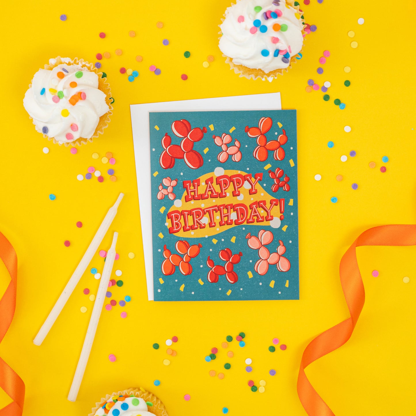 A turquoise birthday card features red, orange and pink balloon dogs, yellow confetti, and the words Happy Birthday! in midcentury-inspired hand lettering. It is on yellow with cupcakes and confetti.
