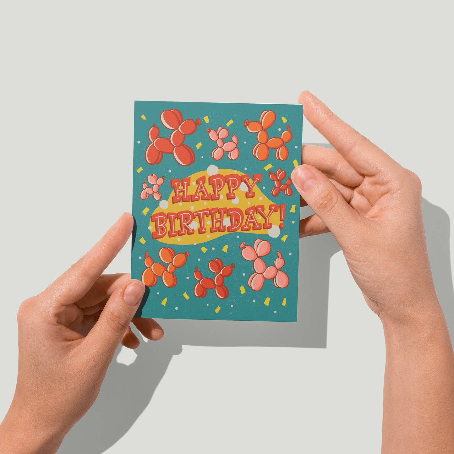 A turquoise birthday card features red, orange and pink balloon dogs, yellow confetti, and the words Happy Birthday in midcentury-inspired hand lettering. A pair of hands holds the card up in front of the camera.
