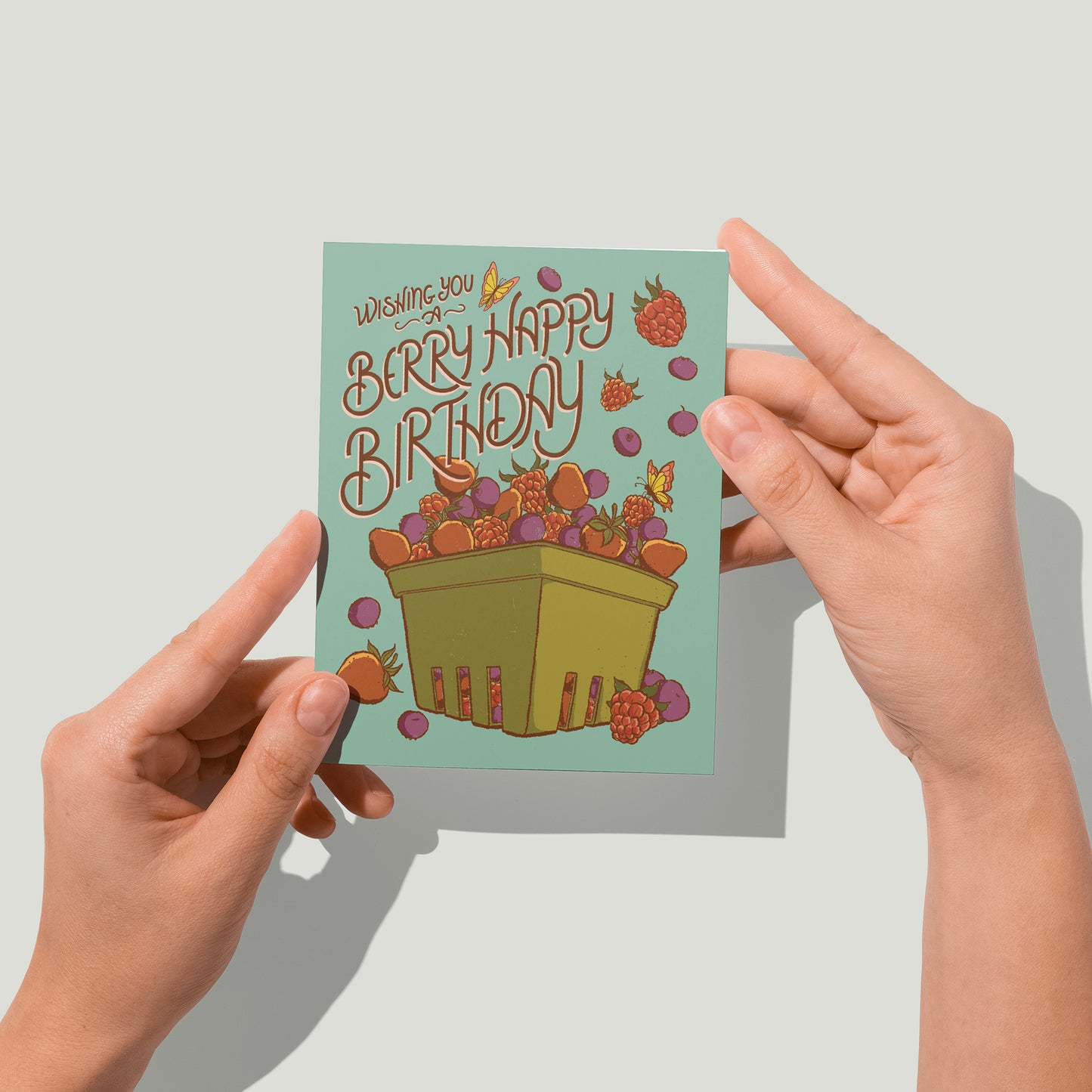A blue greeting card is covered in rustic-looking strawberries, raspberries and blueberries in a green berry basket and reads Have a Berry Happy Birthday. A pair of hands holds the card up in front of the camera.