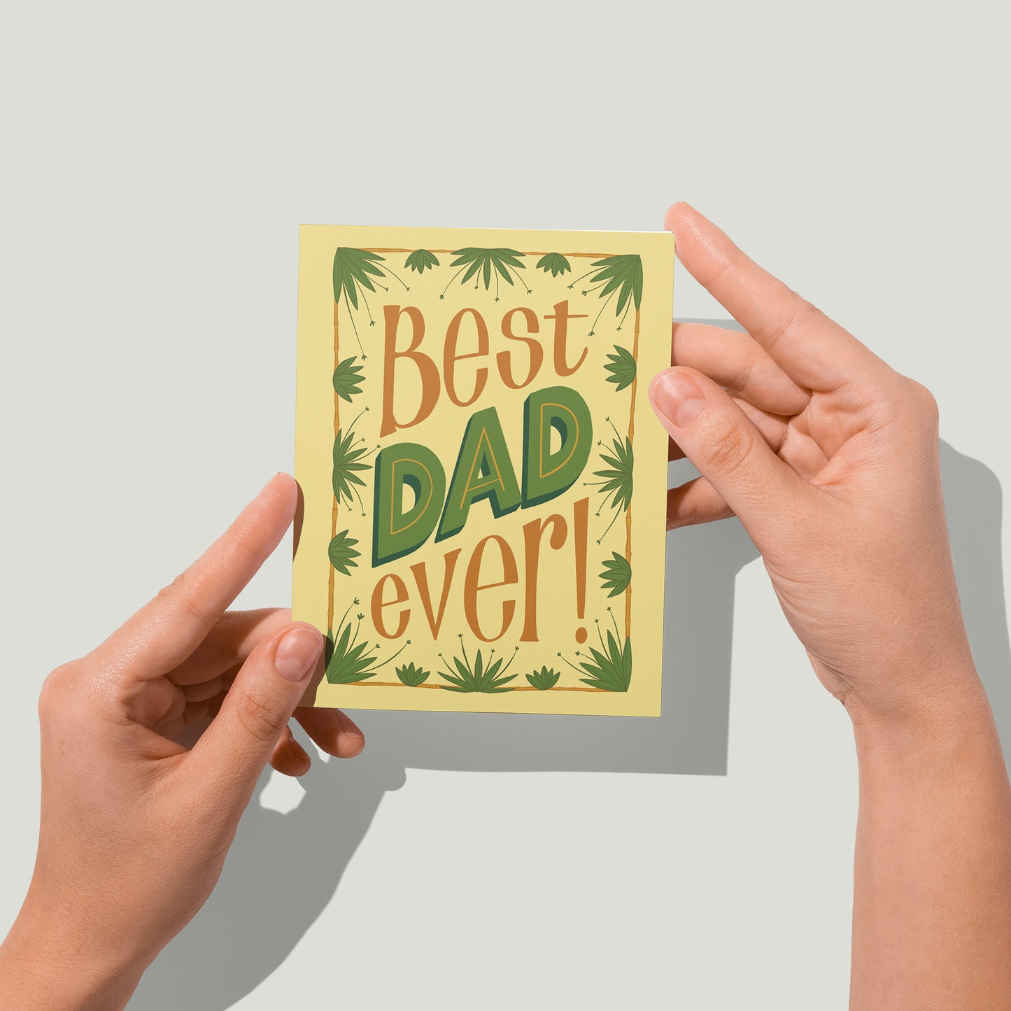 A pale yellow father's day greeting card reads Best DAD ever in brown and green hand lettering. The letters are framed in a brown rectangle out of which sprout green tropical plants. A pair of hands holds the card up in front of the camera.