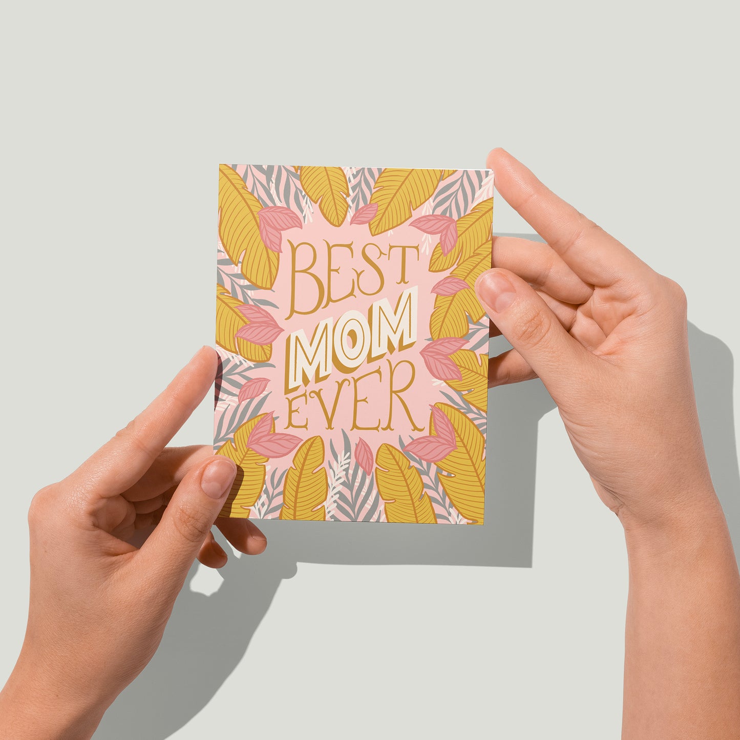 A pink greeting card is covered in golden yellow, white and grey tropical leaves along with the lettered phrase Best Mom Ever in block letters. A pair of hands holds the card up in front of the camera.