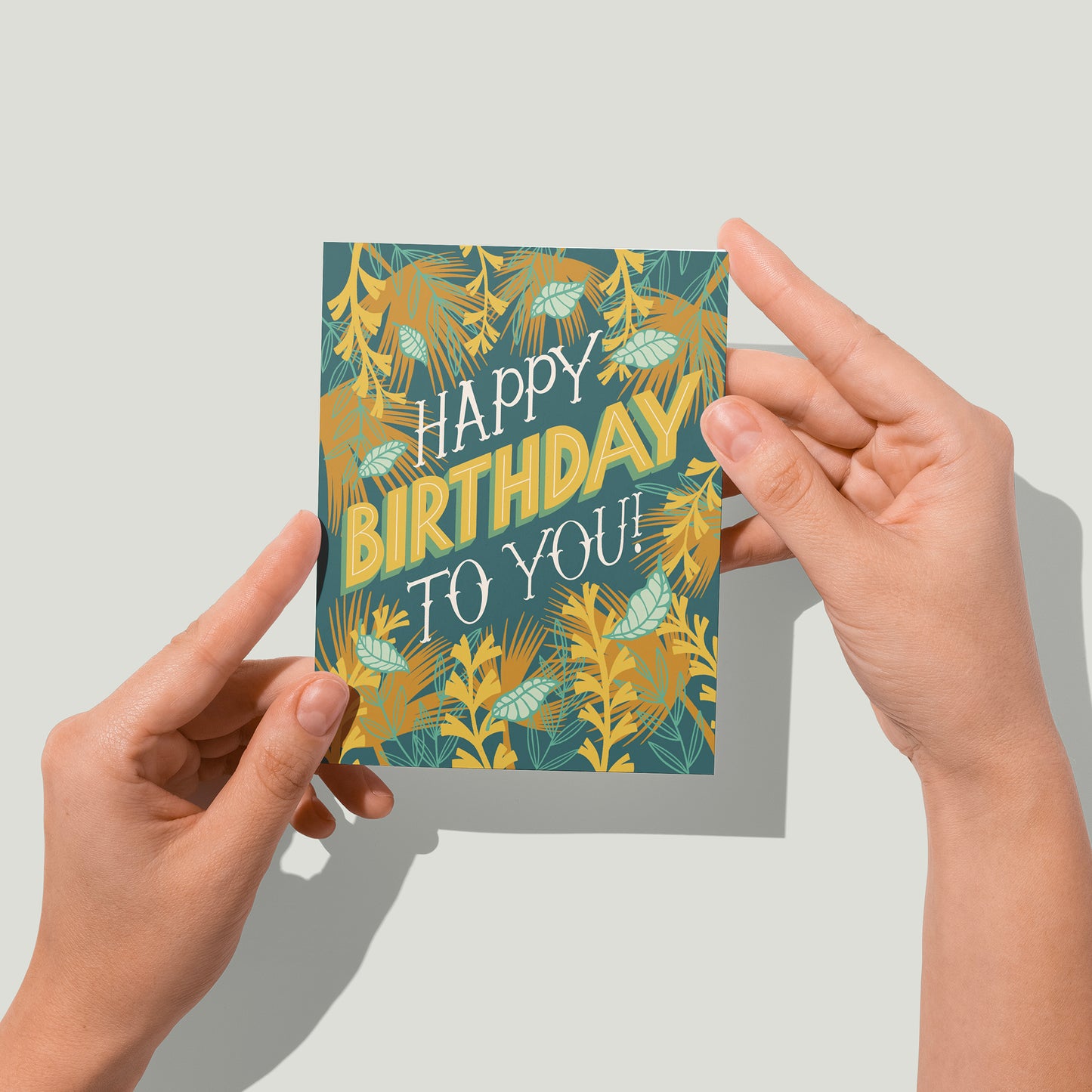 A dark blue greeting card features the hand-lettered words Happy Birthday to you!. The letters are framed by illustrations of tropical leaves and vines. A pair of hands holds the card up in front of the camera.