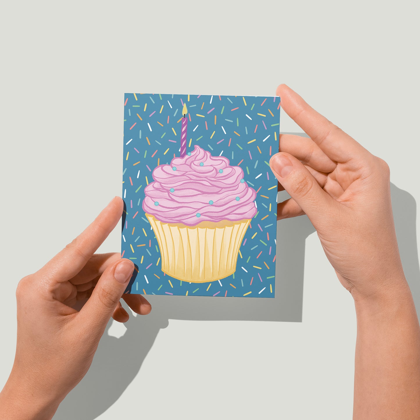 A blue greeting card features a shower of rainbow sprinkles and a cupcake in a paper wrapper topped with swathes of pink frosting and round blue sprinkles. A lit birthday candle is plunked into the frosting. A pair of hands holds the card up in front of the camera.