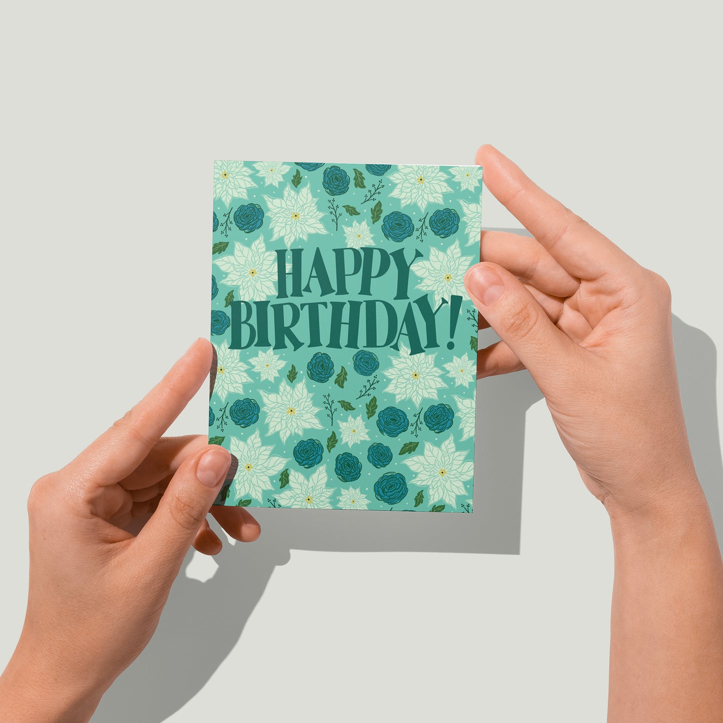 A blue greeting card featuring the words Happy Birthday! in midcentury-inspired dark blue capital letters. Behind and all around the letters are white poinsettias, blue ranunculus flowers, green leaves and black branches. A pair of hands holds the card up in front of the camera.