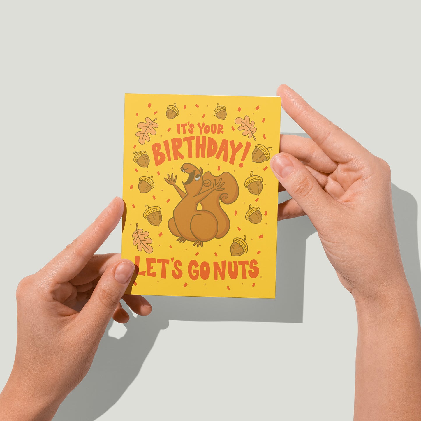 A yellow greeting card shows a joyful squirrel amidst the orange hand-lettered words, It's your birthday! Let's go nuts. The squirrel is showered by acorns, oak leaves, and orange confetti. The card sits against a white envelope on a white background. A pair of hands holds the card up in front of the camera.