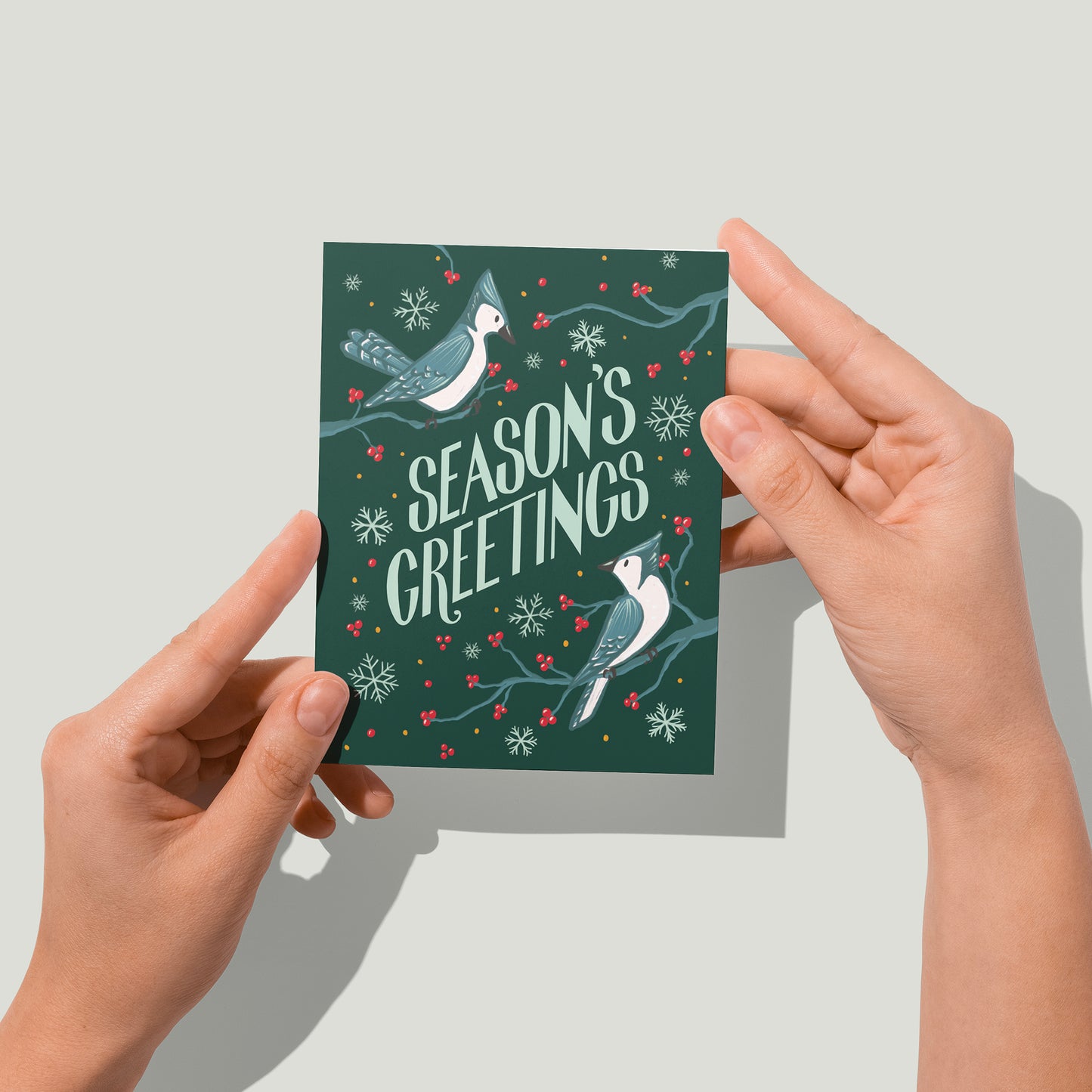 A dark green holiday card shows the words Season's Greetings in all capital letters. Around the letters are two blue jays perched on branches with red berries, along with some loose red berries and snowflakes. A pair of hands holds the card up in front of the camera.