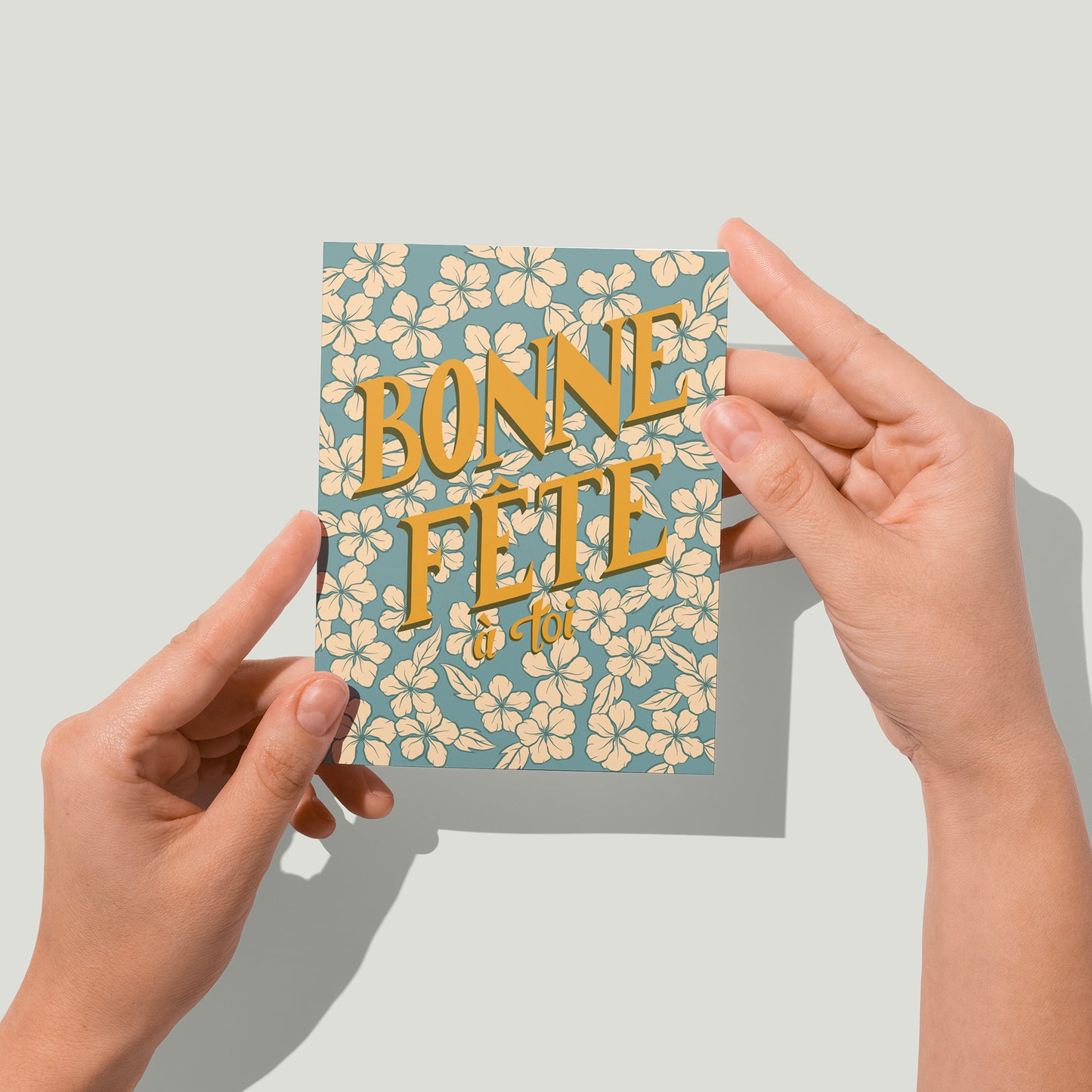 A retro-looking blue greeting card shows creamy flowers and hand lettering that spells out Bonne Fête à toi. A pair of hands holds the card up to the camera.