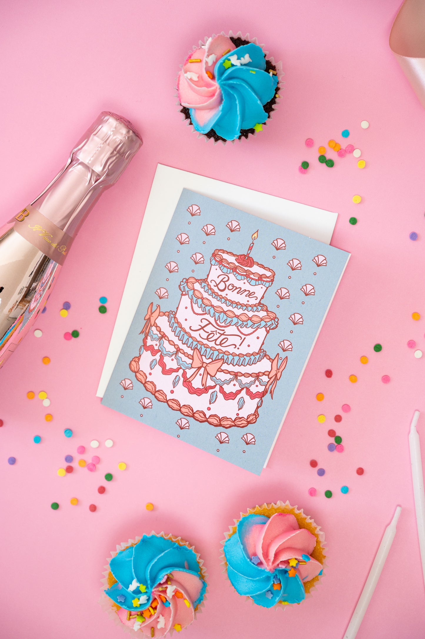 A greeting card shows a rococo birthday cake with the words bonne fête and it sits on a pink background with confetti, candles and cupcakes.