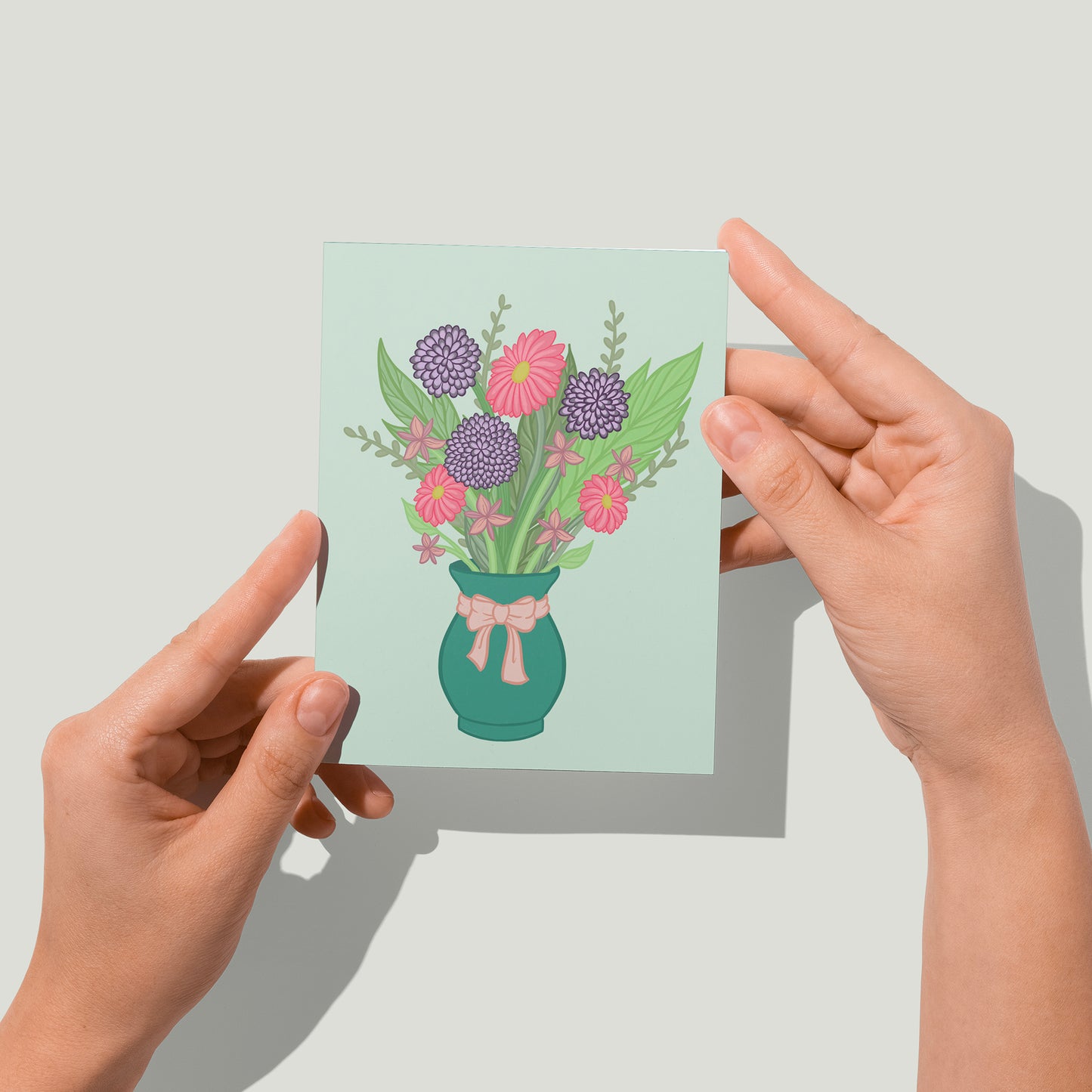 A pastel mint greeting card features an illustration of a bouquet in a green vase with a pink bow around it. The bouquet includes purple and pink flowers and greenery in various shades. A pair of hands holds the card up in front of the camera.