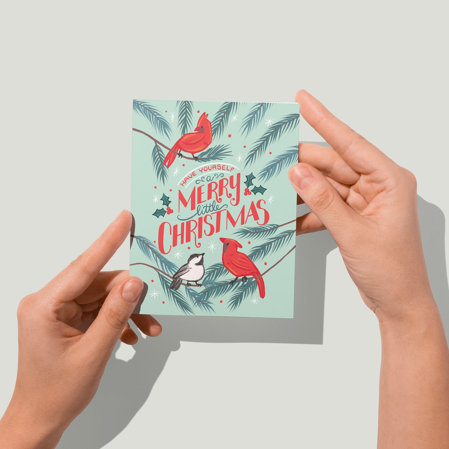A greeting card with the words Have Yourself a Merry Little Christmas lettered in red and green, surrounded by red cardinals and a chickadee on a mint background. A pair of hands holds the card up in front of the camera.