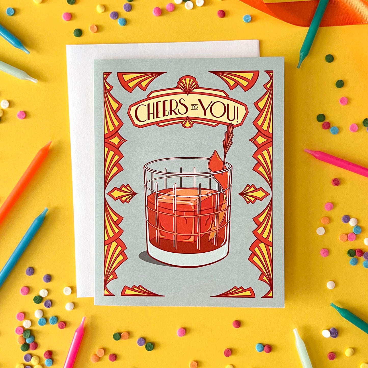A greeting card says 'cheers to you' in art deco lettering above an old fashioned. The card is on yellow with confetti and candles.