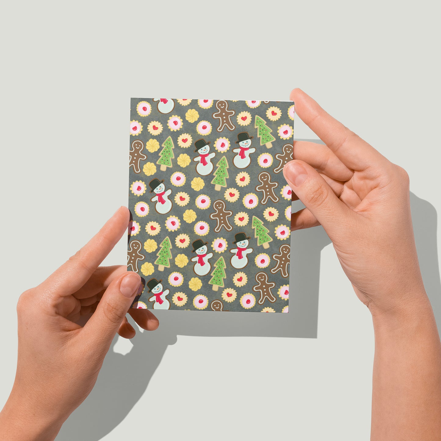 A greeting card is patterned with a variety of Christmas cookies - gingerbread men, snowmen, sugar cookie trees, empire cookies and shortbread. A pair of hands holds the card up in front of the camera.