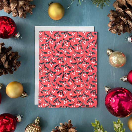 A red greeting card shows a blue and white botanical mistletoe pattern. The card is surrounded by red and gold ornaments, pinecones, and greenery.
