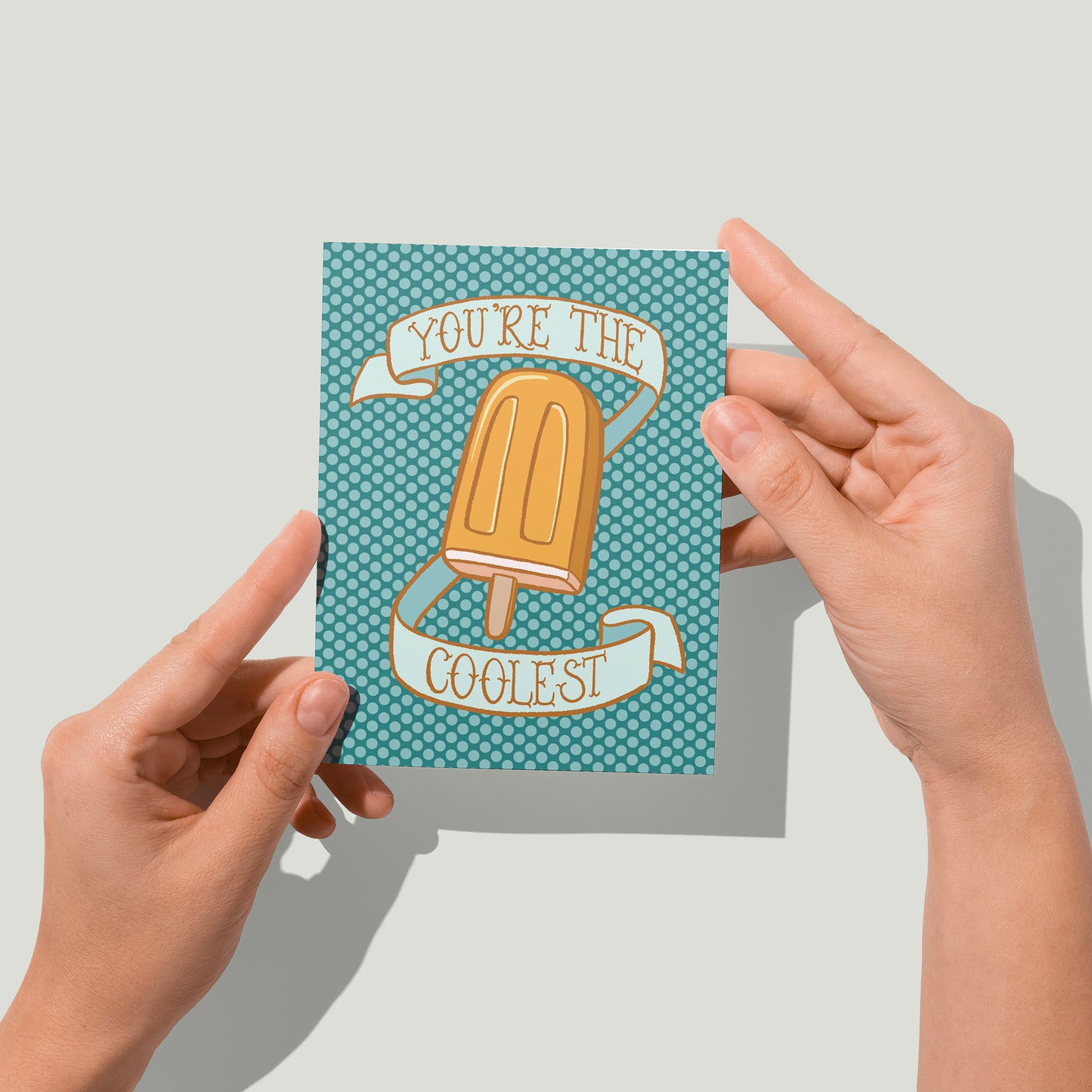 A turquoise polka dot greeting card shows an illustration of a creamy orange popsicle surrounded by a banner that reads you're the coolest in hand lettering. A pair of hands holds the card up in front of the camera.