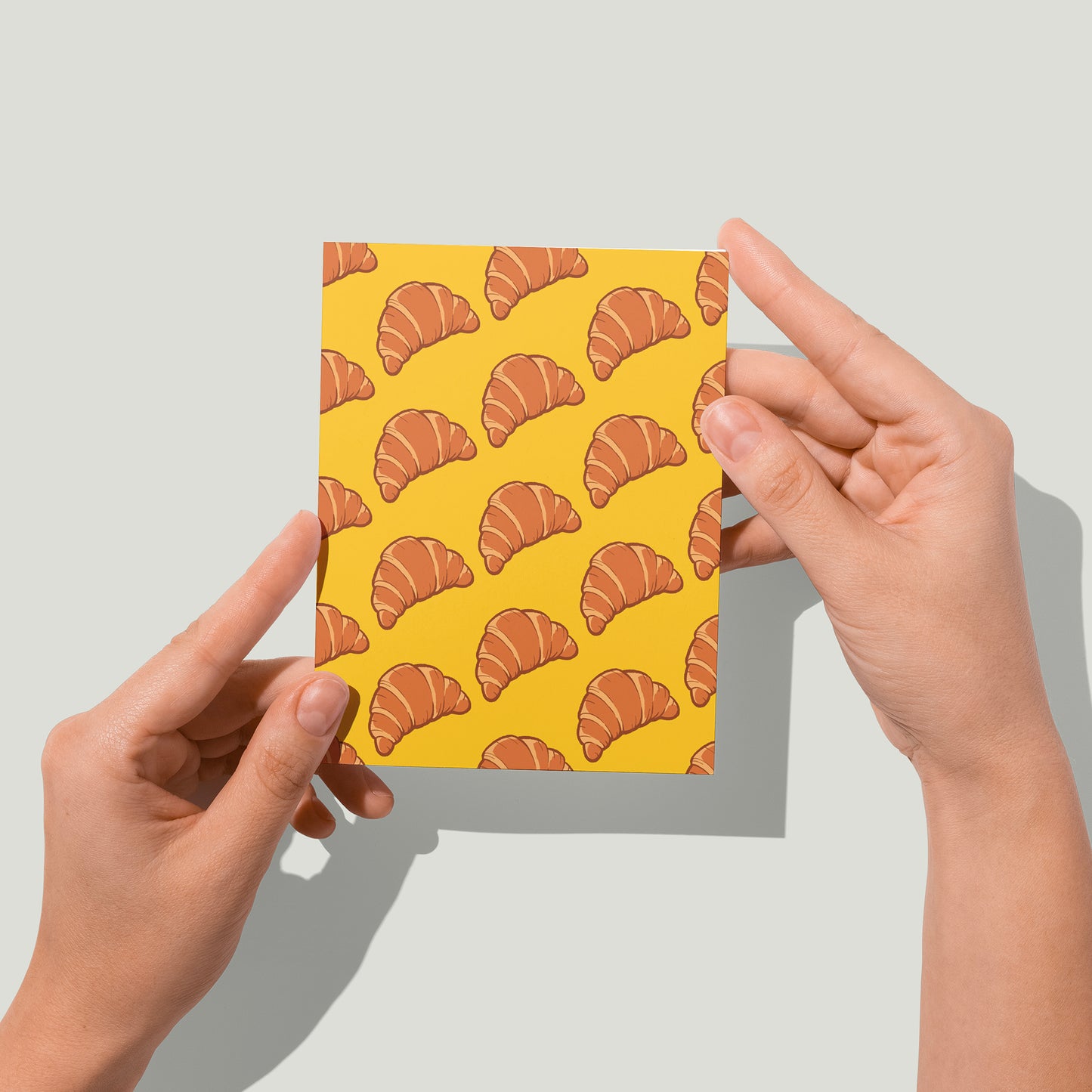 A yellow greeting card is patterned with drawings of croissants in diagonal lines. A pair of hands holds the card up in front of the camera.