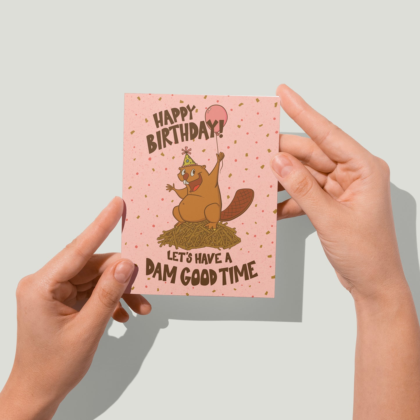 A pink birthday card says Happy Birthday! Let's Have a Dam Good Time in hand-lettering. On the card is a beaver in a party hat on a dam. A pair of hands holds the card up to the camera.