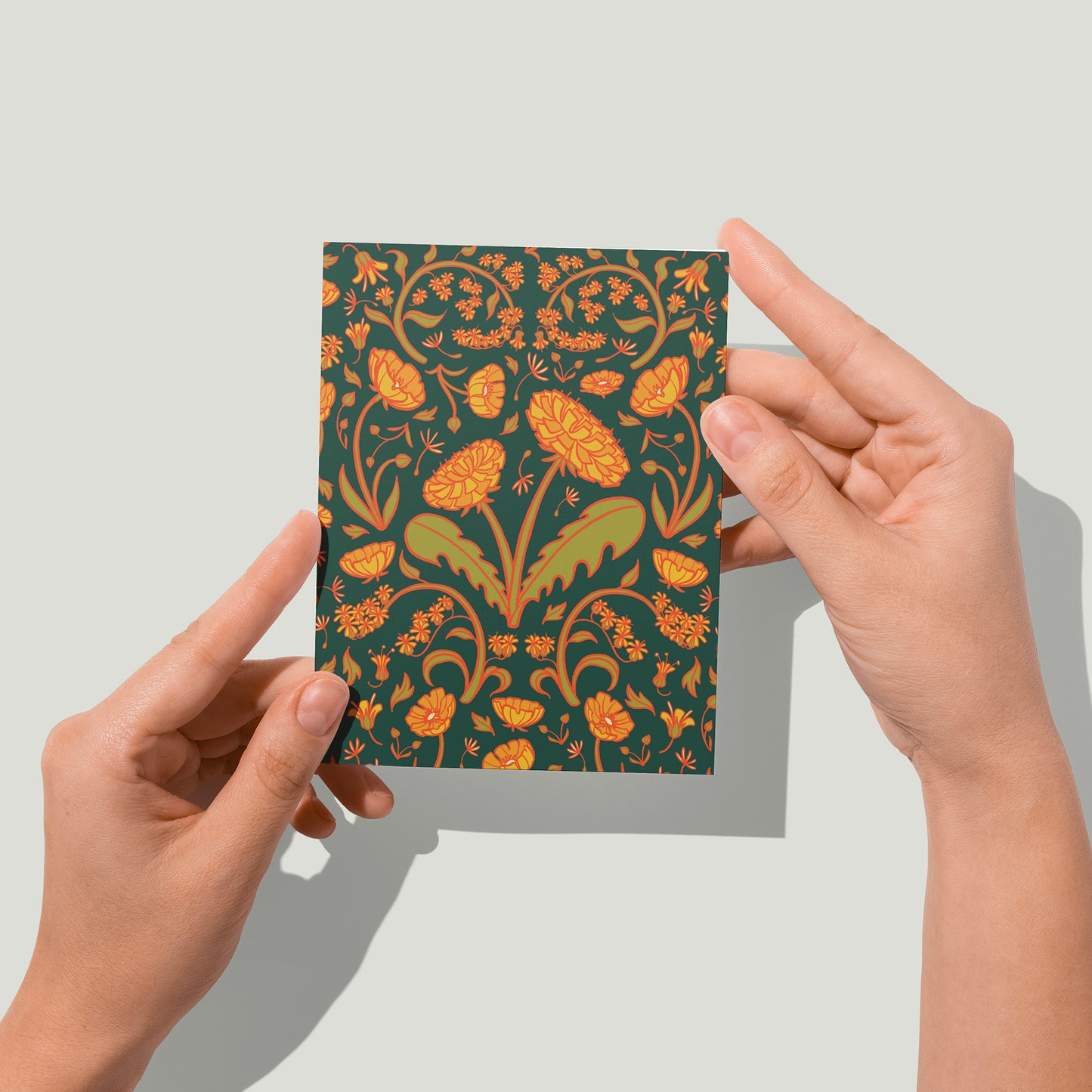 A dark green card is covered in a floral pattern of dandelions, buttercups and goldenrod. A pair of hands holds the card up in front of the camera.