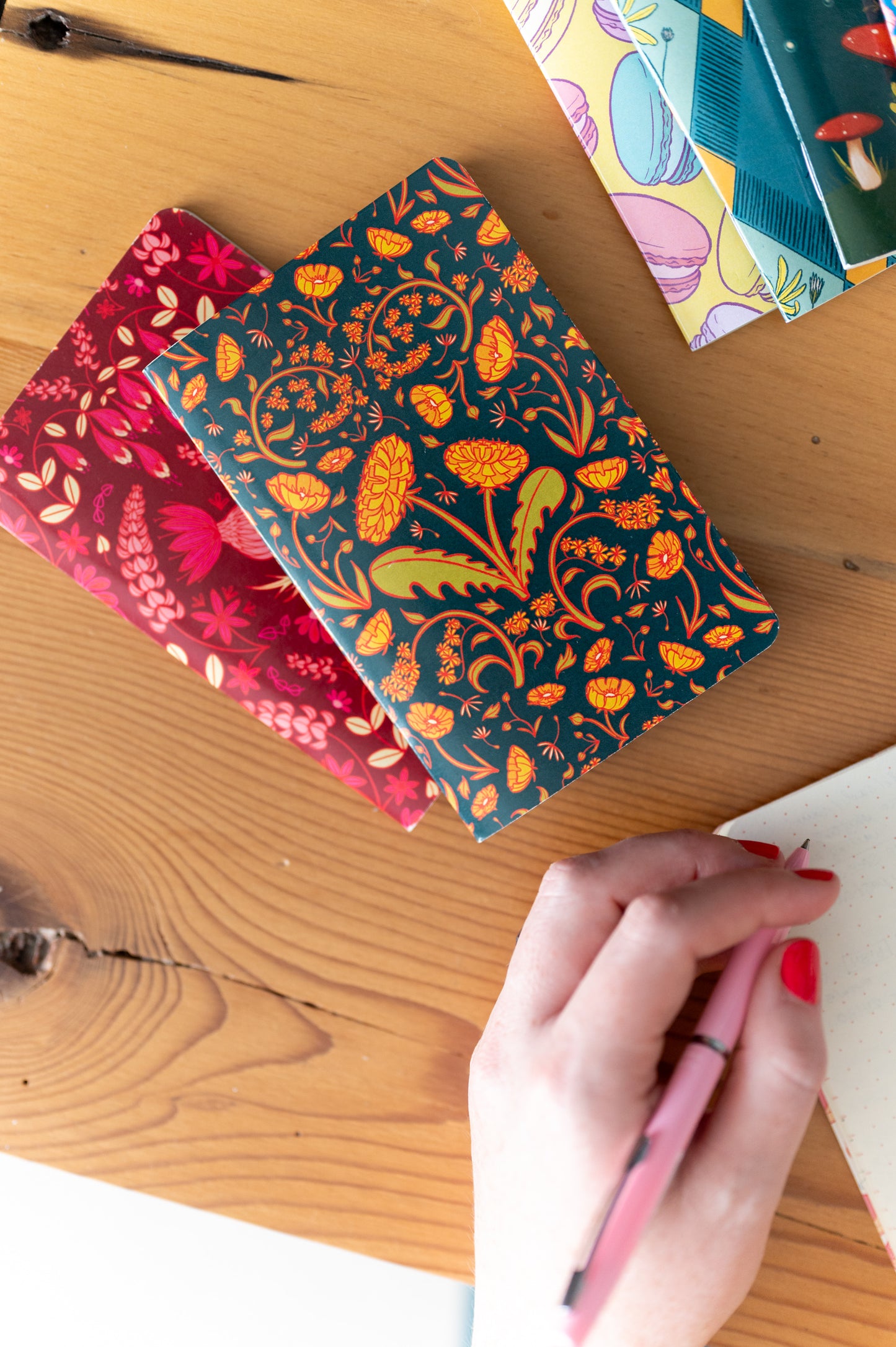 A dot grid pocket notebook is open and a person holds a pen above it. Above it are other pocket notebooks, two with floral covers.