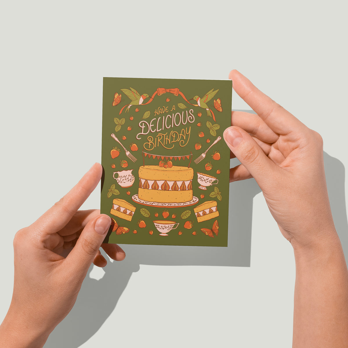 An olive green greeting card features hummingbirds, teacups, butterflies, strawberries, and cake, along with the words Have a Delicious Birthday in hand-lettering. A pair of hands holds the card up in front of the camera.