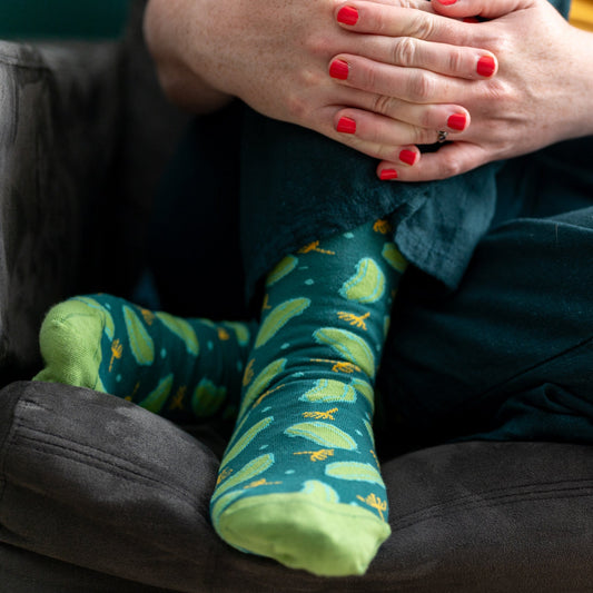 A person wears a pair of green dill pickle patterned socks. They sit on a grey armchair.