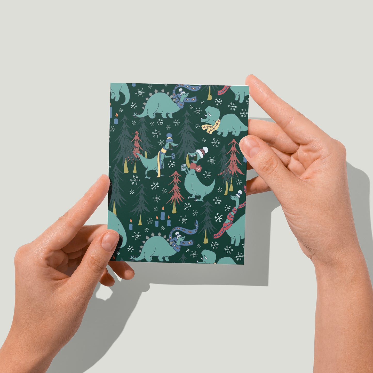 A greeting card is covered with a pattern of dinosaurs in toques and scarves with mugs of hot chocolate. Scattered among the dinosaurs are trees and candles. A pair of hands holds the card up in front of the camera.
