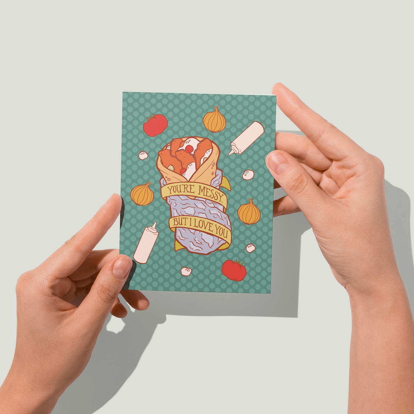 A green polka dot greeting card features an illustration of a donair wrapped in foil, with squeeze bottles, onions and tomatoes all around it and a banner that reads You're Messy But I Love You. A pair of hands holds the card up in front of the camera.