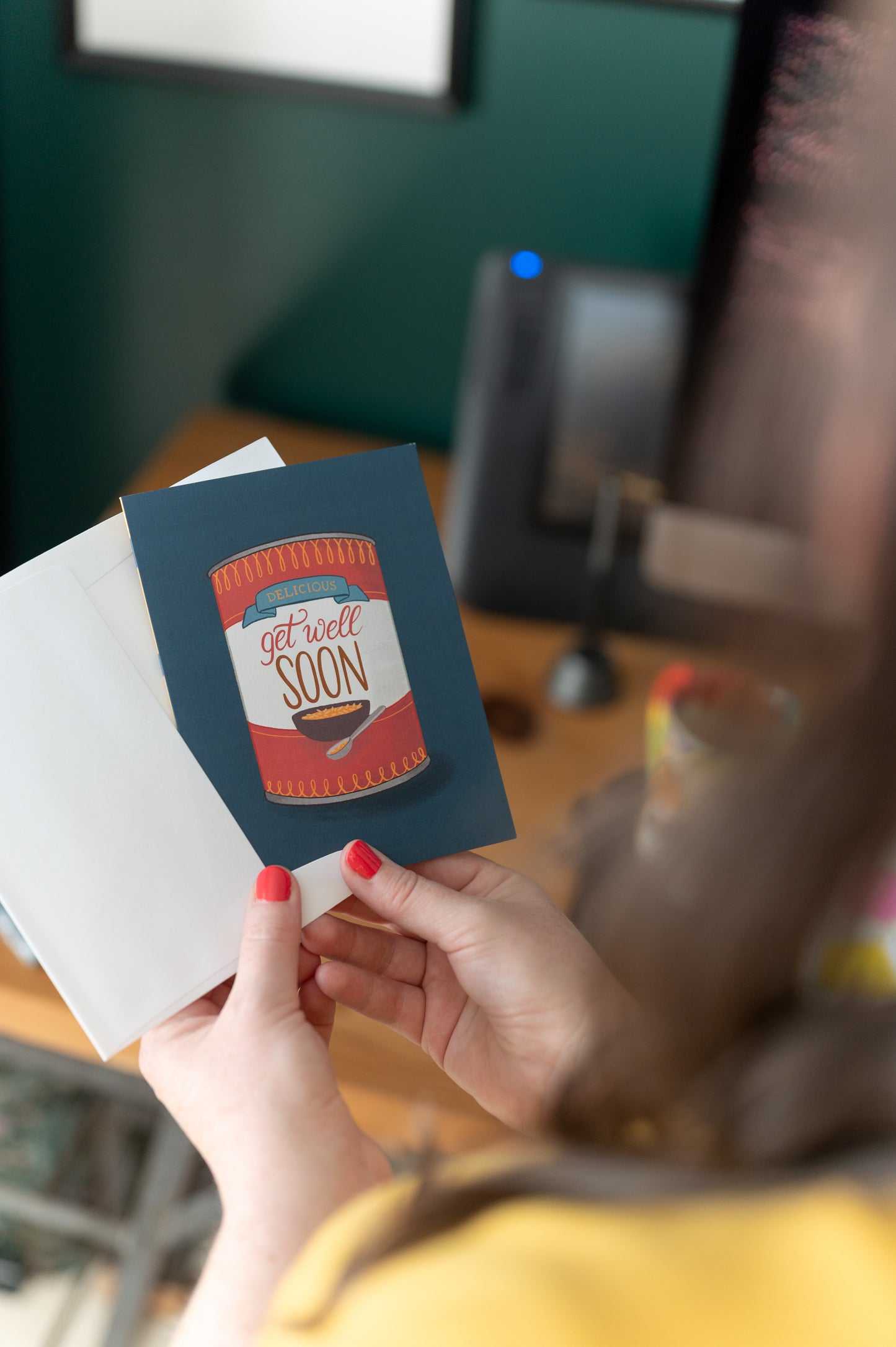 A person holds a get well soon greeting card with a soup can on it in a white envelope.