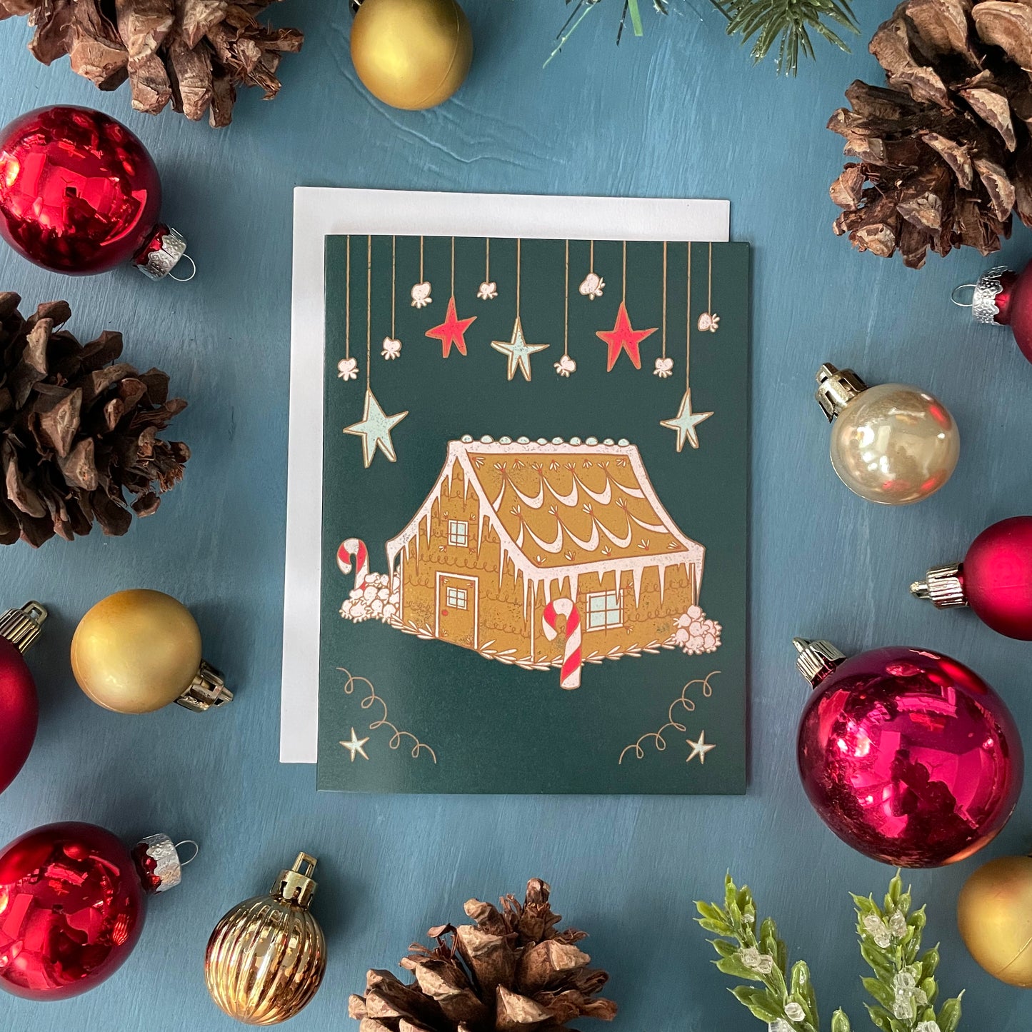 A dark green greeting card features hanging stars and a frosted gingerbread house among popcorn snowbanks. The card is among red and gold ornaments, pinecones, and greenery on a blue background.
