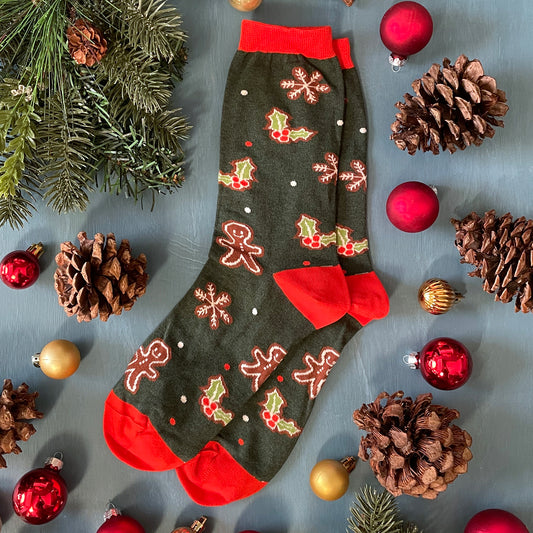 A pair of dark green socks patterned with gingerbread people, snowflakes and holly lie flat. They are surrounded by pinecones and holiday ornaments.