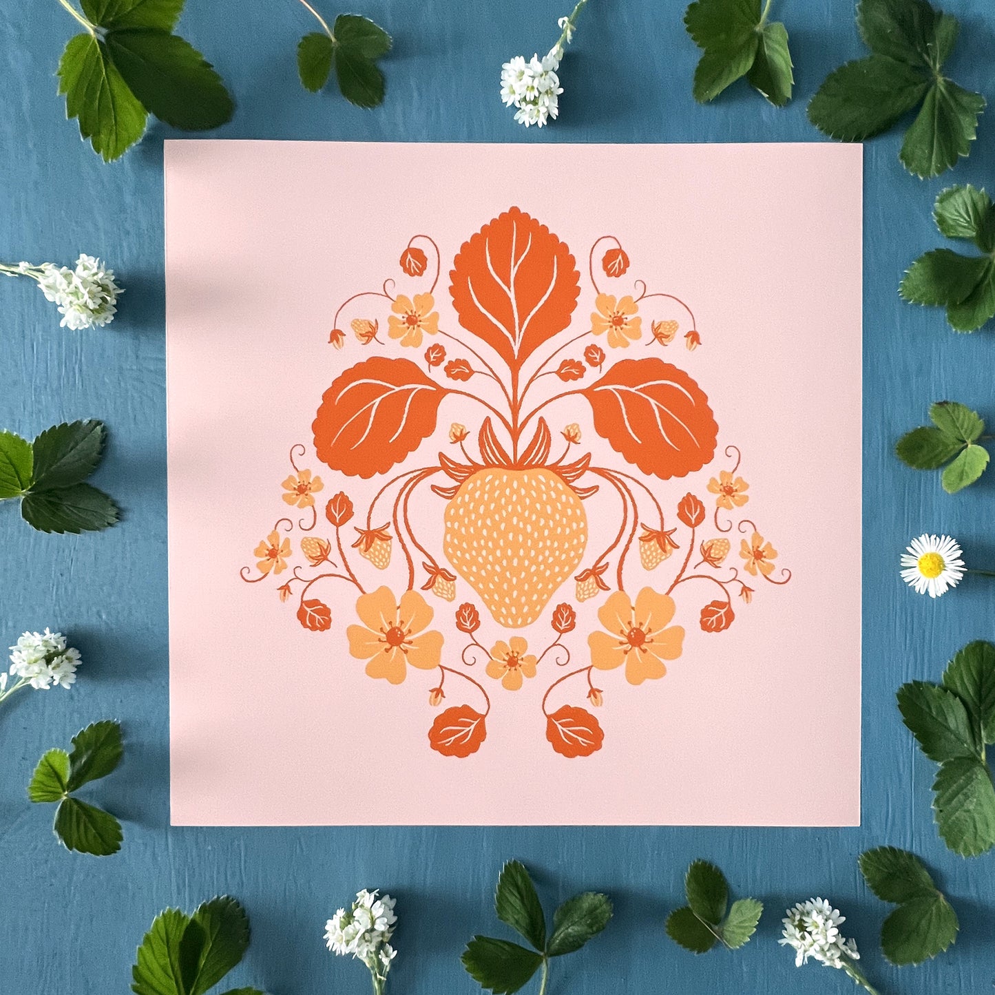 A pink and orange square botanical art print of a strawberry along with leaves and strawberry blossoms is centred on a blue background surrounded by wildflowers and wild strawberry leaves.