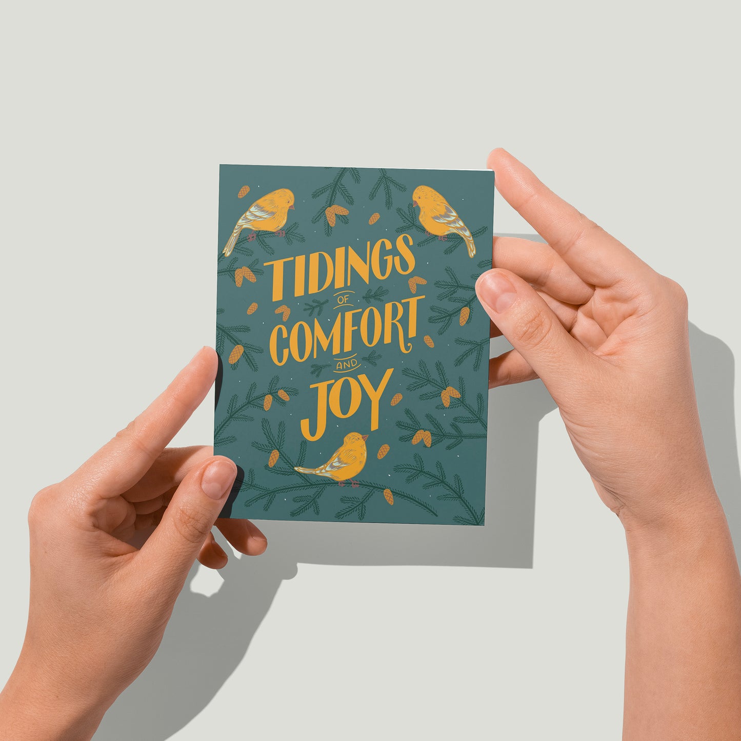 A greeting card features yellow hand-lettering reading tidings of comfort and joy on a blue background. There are painted goldfinches and pinecone-laden branches around the words. A pair of hands holds the card up in front of the camera.