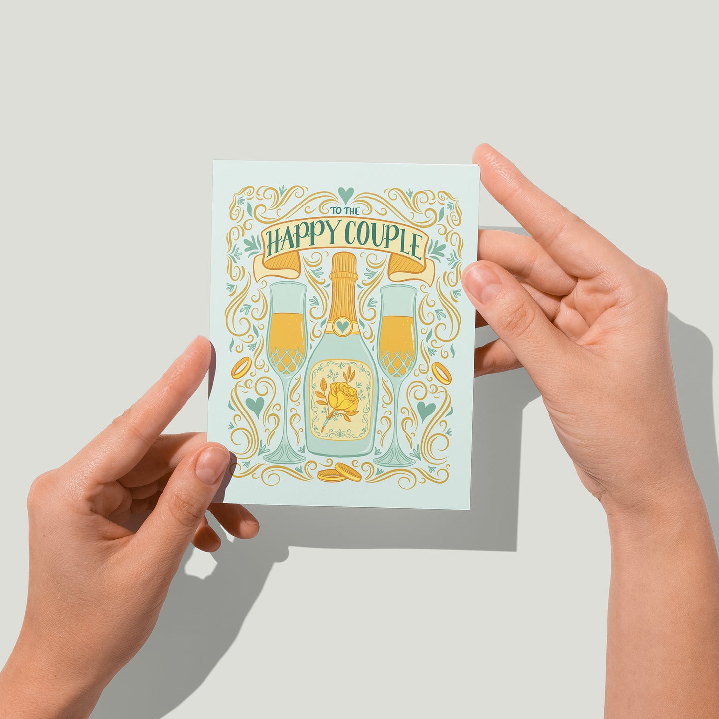 A pastel blue greeting card features an illustration of two full champagne flutes, a champagne bottle with a yellow rose, wedding bands, Art Nouveau flourishes, hearts, and the words To The Happy Couple. A pair of hands holds the card up in front of the camera.