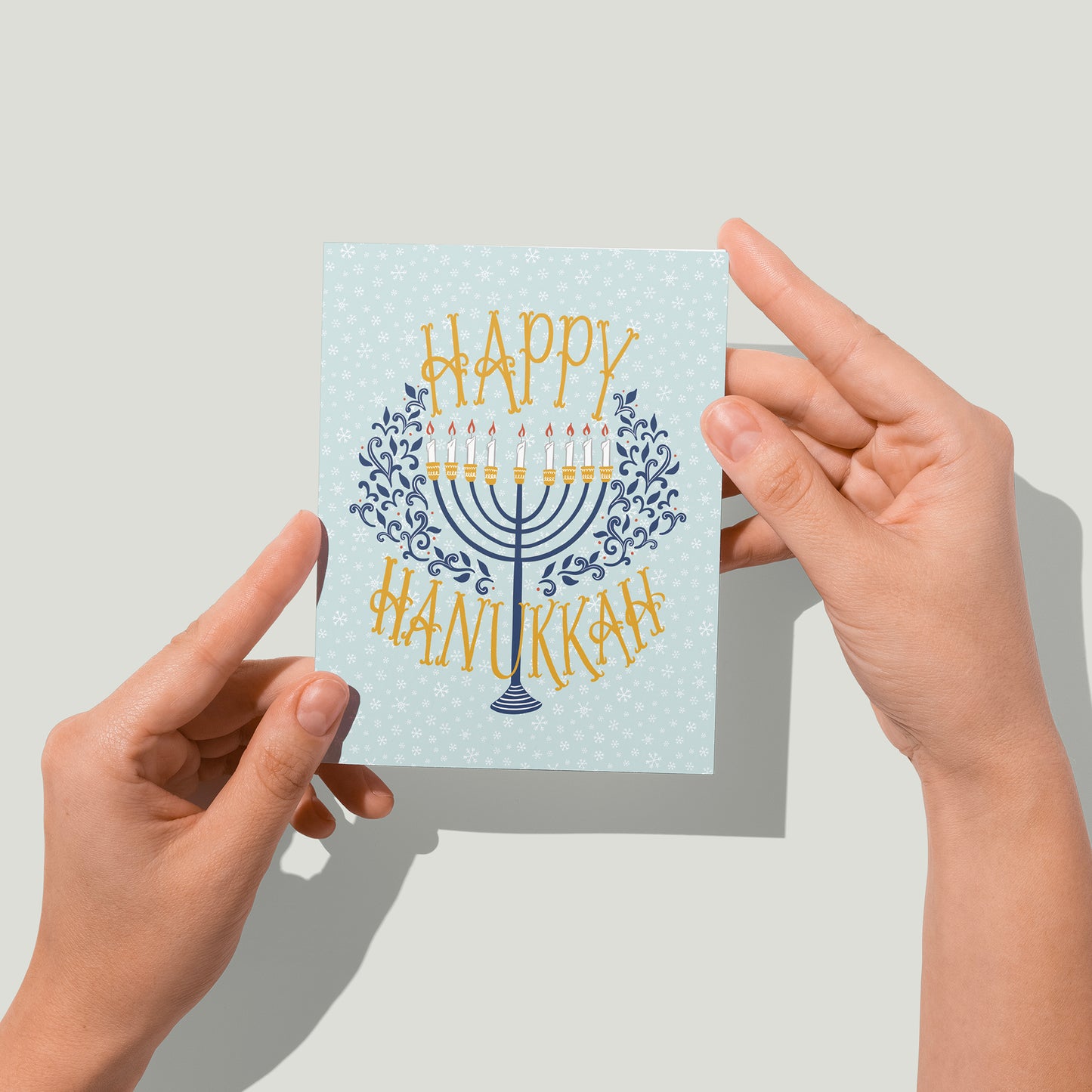 A pale blue snowflake-patterned greeting card reads Happy Hanukkah in deep yellow hand-lettering. In between the words is a navy menorah with lit candles flanked by navy flourishes. A pair of hands holds the card up in front of the camera.