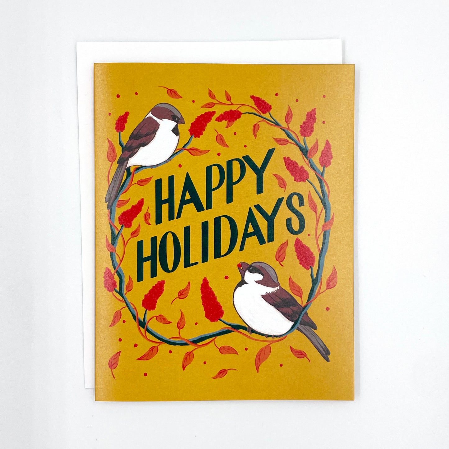 A yellow greeting card features the words Happy Holidays in hand lettering, surrounded by sparrows and staghorn sumac. The card is surrounded by red and gold ornaments, pinecones, and greenery. The card is on a white envelope on a white background.