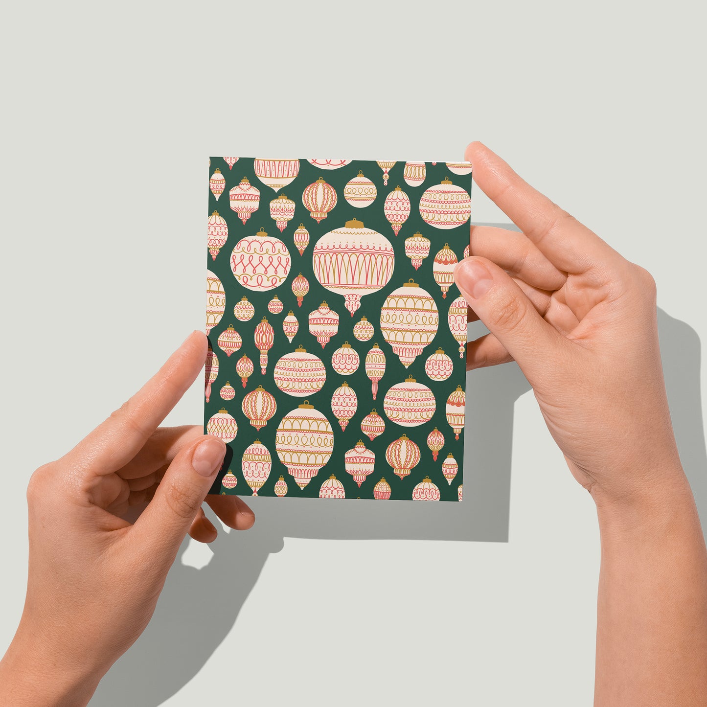 A green greeting card boasts a pattern of cream, red and gold tree ornaments. A pair of hands holds the card up in front of the camera.