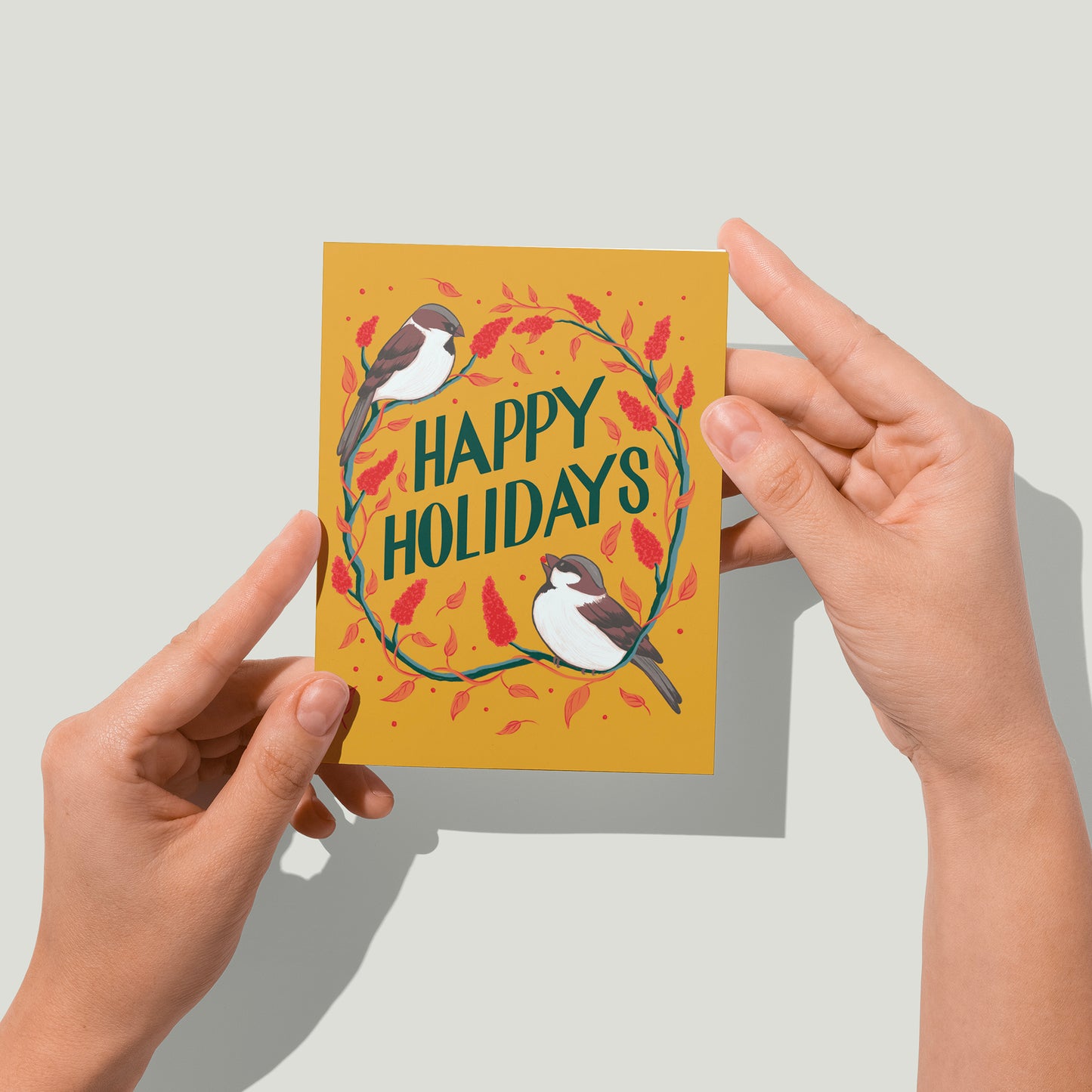 A yellow greeting card features the words Happy Holidays in hand lettering, surrounded by sparrows and staghorn sumac. The card is surrounded by red and gold ornaments, pinecones, and greenery. A pair of hands holds the card up in front of the camera.