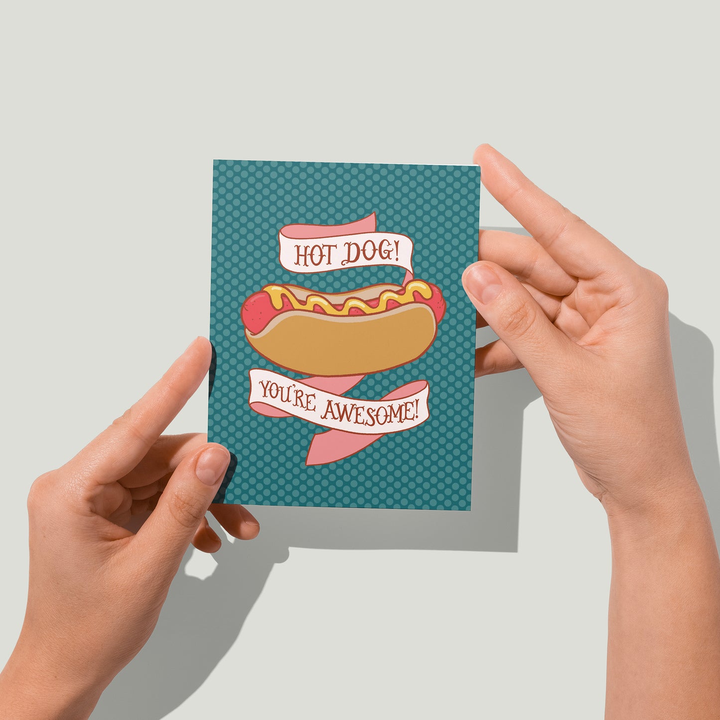 A greeting card is deep blue-green and features an illustration of a hot dog on a polka dot background. Behind the hot dog is a banner that reads Hot Dog! You're Awesome! in hand lettering. A pair of hands holds the card up in front of the camera.