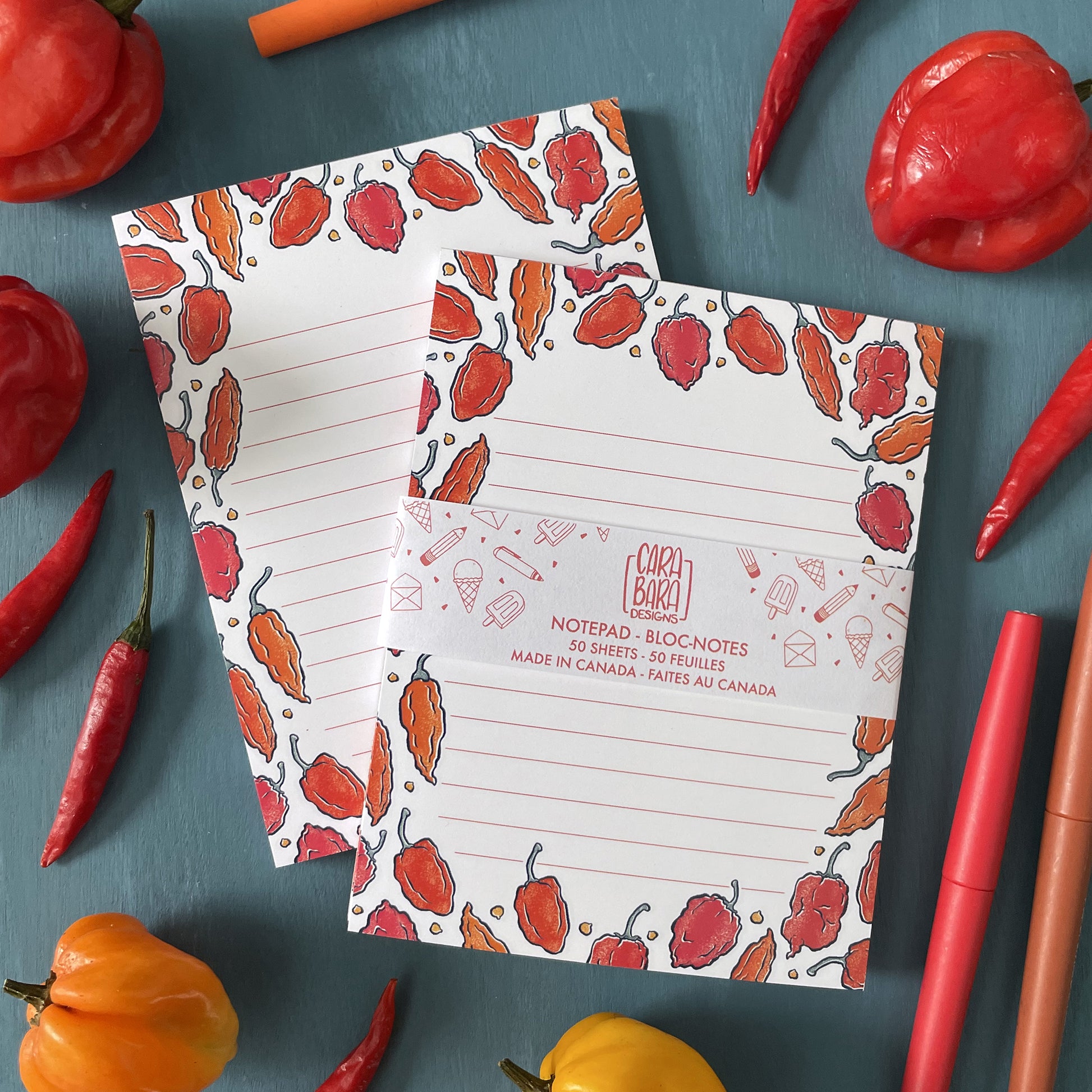 A pair of white lined notepad features illustrations of hot peppers in orange and red. The front notepad is packaged in a belly band with the Carabara Designs logo and says the notepad is 50 pages and made in Canada. The tear-off notepads are surrounded by orange and red Scotch bonnet peppers, red thai chiles, and a red pen.