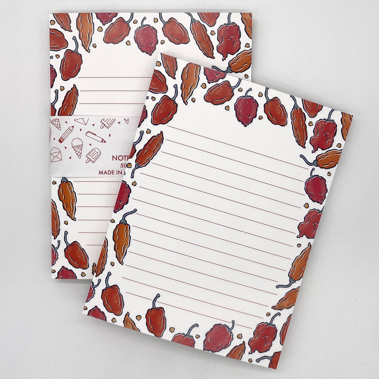 A pair of white lined notepads feature illustrations of hot peppers in orange and red. The back notepad is packaged in a belly band indicating that the notepad is 50 pages and made in Canada.