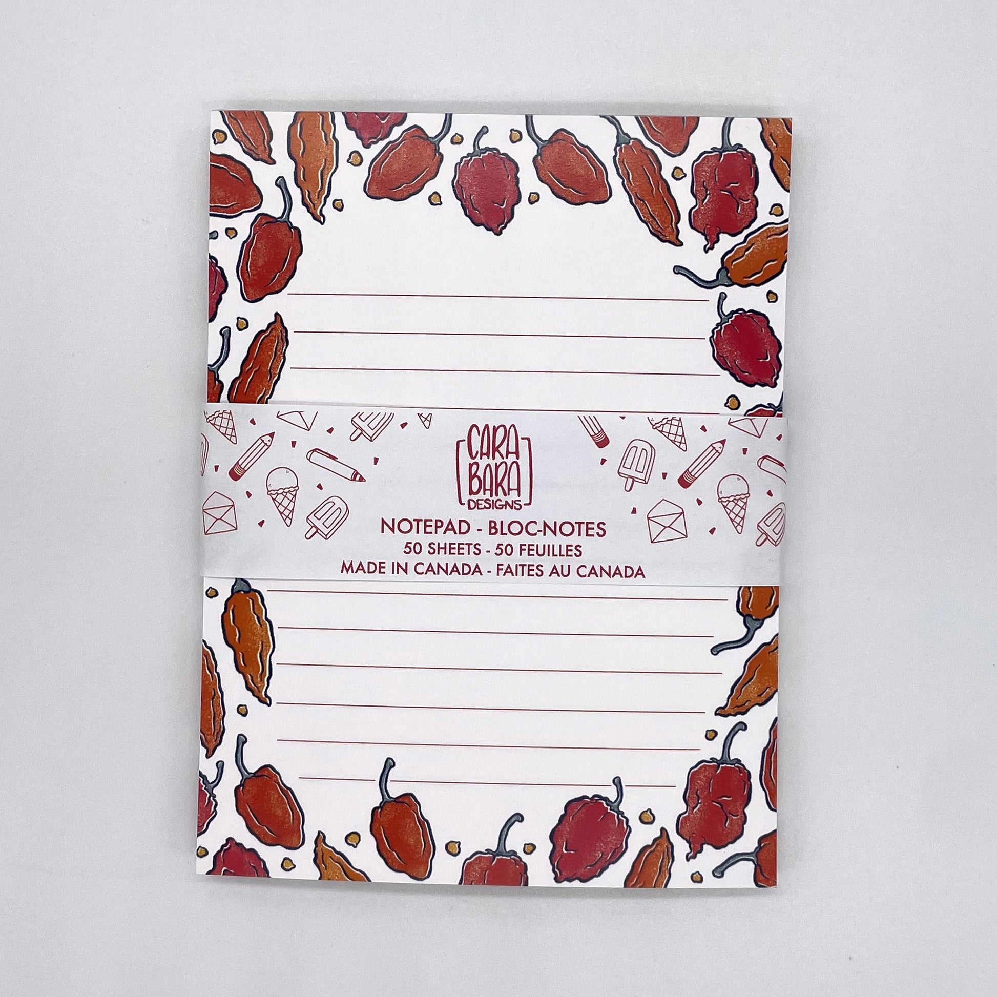 A white lined notepad features illustrations of hot peppers in orange and red. It is packaged in a belly band with the Carabara Designs logo, indicating the notepad is 50 pages and made in Canada.