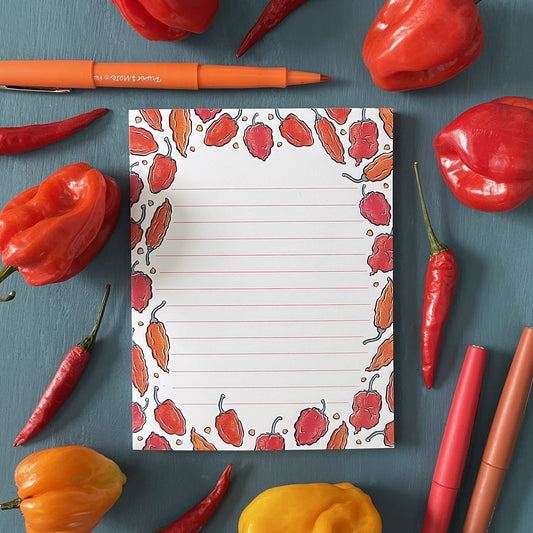 A white lined notepad features illustrations of hot peppers in orange and red. The tear-off notepad is surrounded by orange and red Scotch bonnet peppers, red thai chiles, and a red pen.