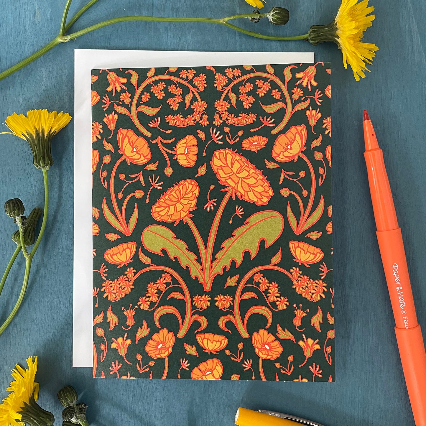 A dark green card is covered in a floral pattern of dandelions, buttercups and goldenrod. It is flanked by yellow flowers and yellow and orange pens.