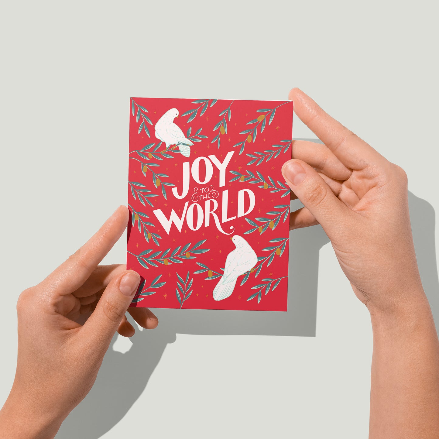 A red greeting card shows two white doves, branches of golden olives, and the hand-lettered words Joy to the World in white. A pair of hands holds the card up in front of the camera.