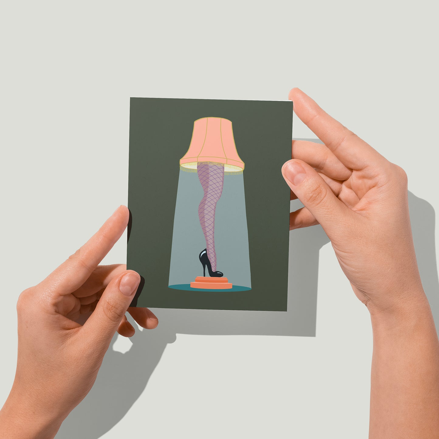 A dark green greeting card features an illustration of a lampshade atop a leg clad in fishnet stockings with a black high heel. A pair of hands holds the card up in front of the camera.
