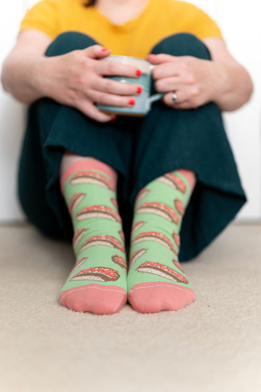 A person wears green and pink socks patterned with lobster rolls. They hold a mug and are leaning against a wall.
