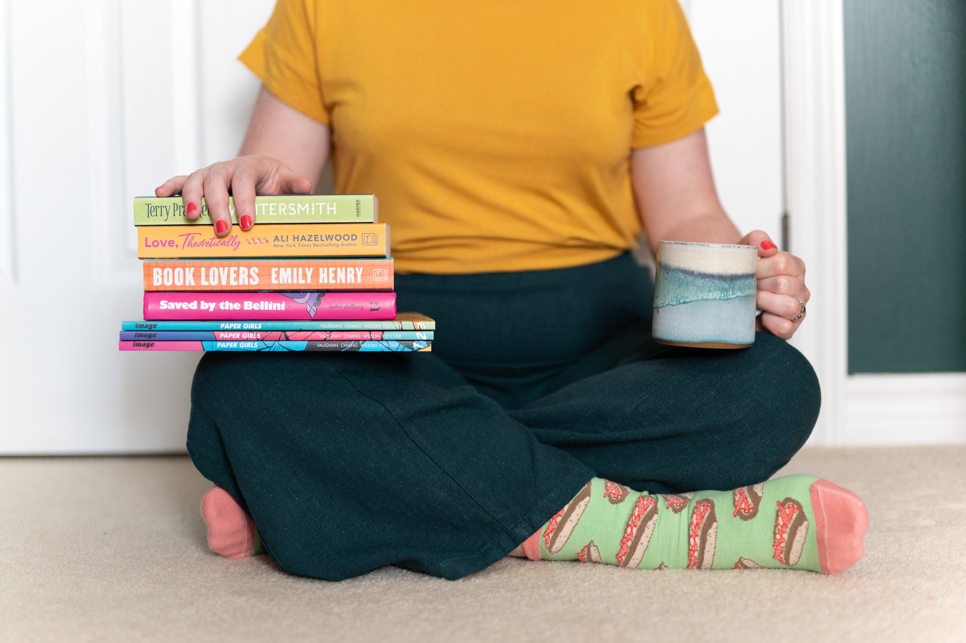 A person wears green and pink socks patterned with lobster rolls. They hold a mug on one knee and a stack of books on the other.