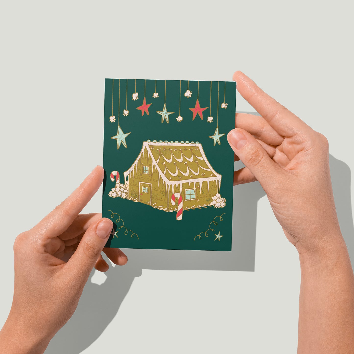 A dark green greeting card features hanging stars and a frosted gingerbread house among popcorn snowbanks. A pair of hands holds the card up in front of the camera.