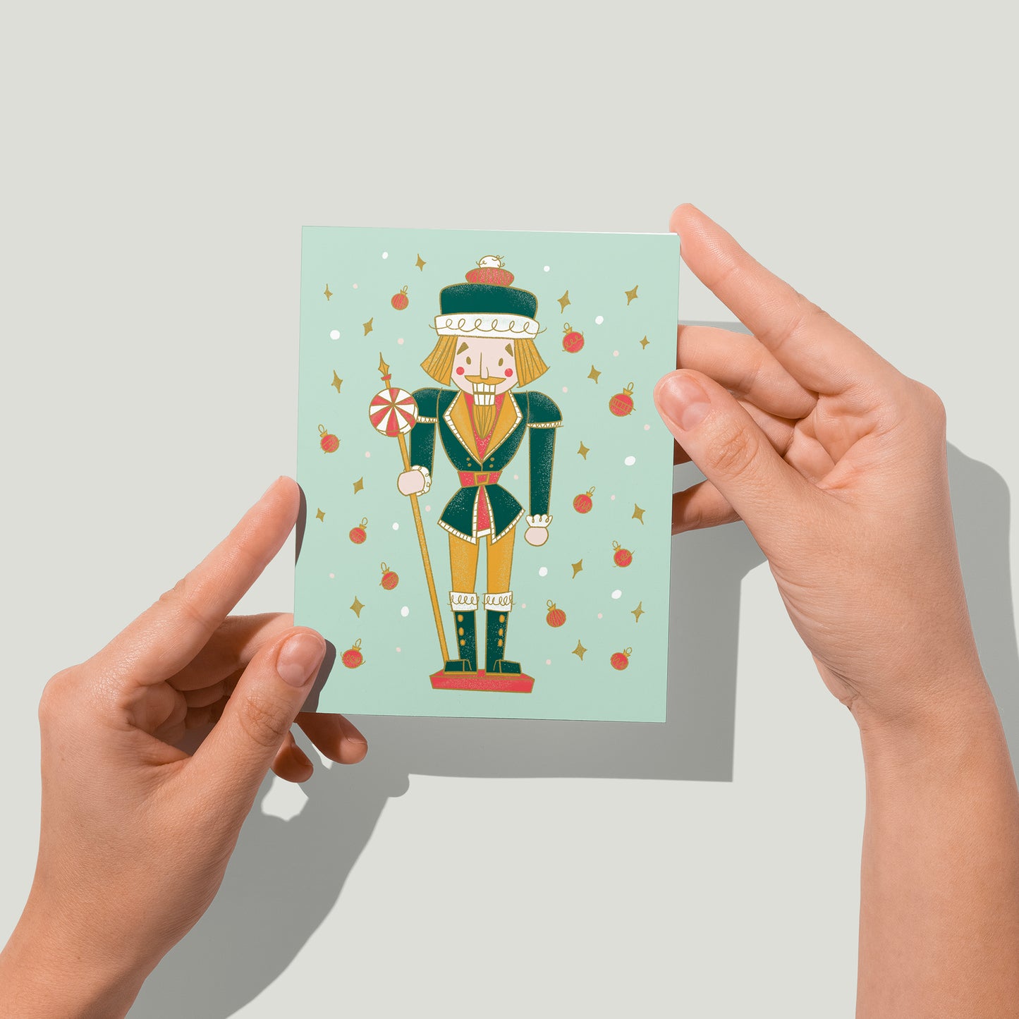 A mint card features a midcentury-style nutcracker in green and yellow, flanked by dots, yellow diamonds and red baubles. A pair of hands holds the card up in front of the camera.