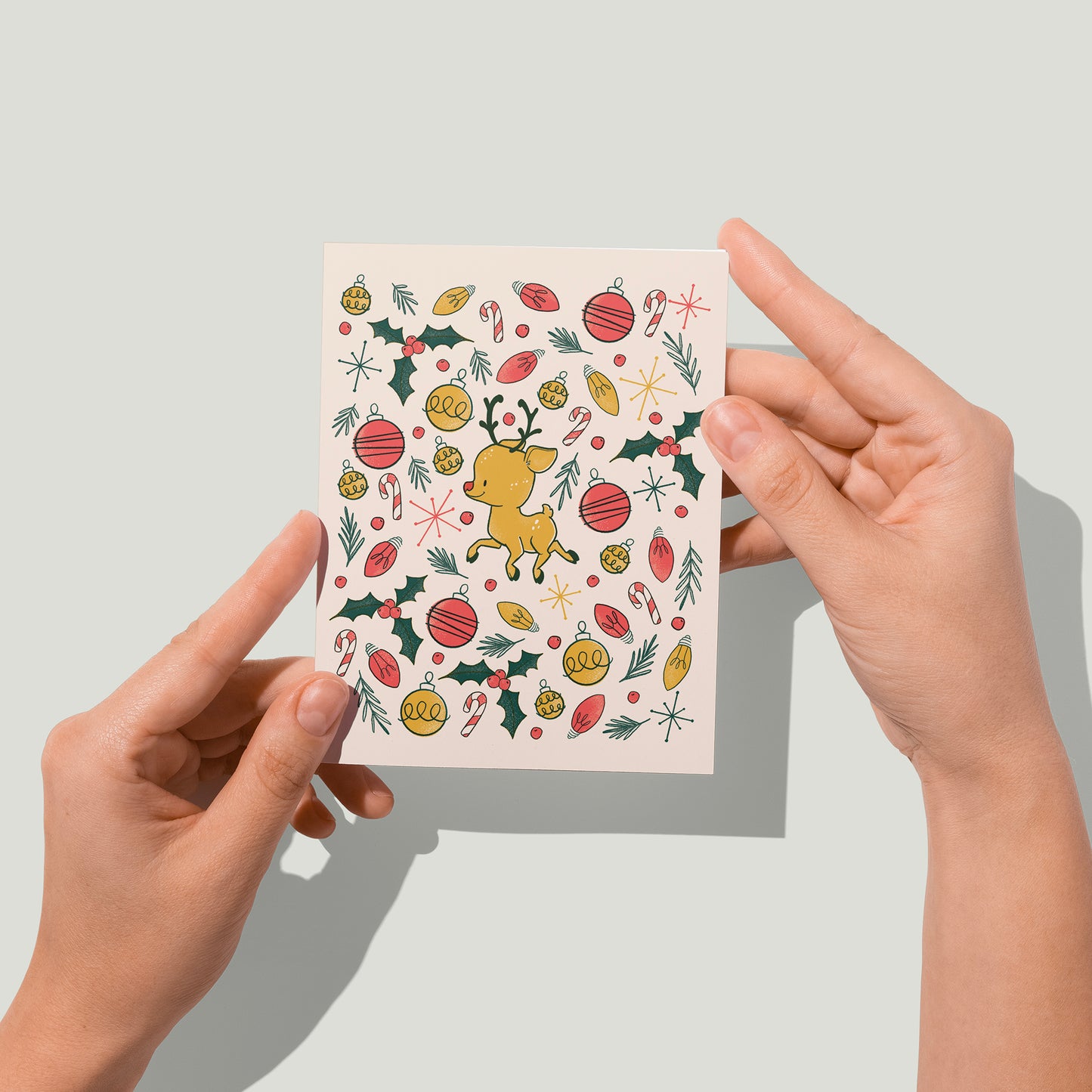 A blush pink card features a cute reindeer surrounded by an explosion of Christmas lights, ornaments, pine branches and holly. A pair of hands holds the card up in front of the camera.